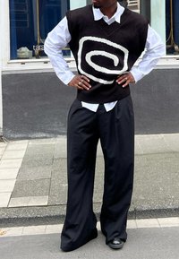Gilet di maglia nera con motivo a spirale bianca, indossato sopra una camicia bianca. Pantaloni neri larghi. Gli accessori includono anelli e orologio.