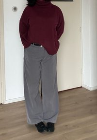 Pull à col roulé bordeaux associé à un pantalon gris à jambes larges. Chaussures noires. Fond uni avec un sol en bois clair.