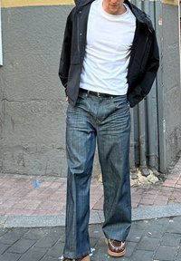 Casaco preto com fecho éclair por cima de uma t-shirt branca, combinado com jeans largos azul-escuro e sapatos castanhos com costura decorativa. Ambiente casual na rua.