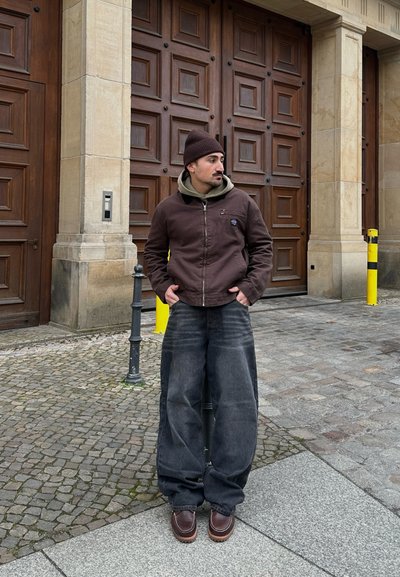 Hombre con un gorro marrón, chaqueta marrón, sudadera verde oliva y vaqueros anchos negros, de pie con las manos en los bolsillos sobre un pavimento de piedra cerca de unas puertas de madera.