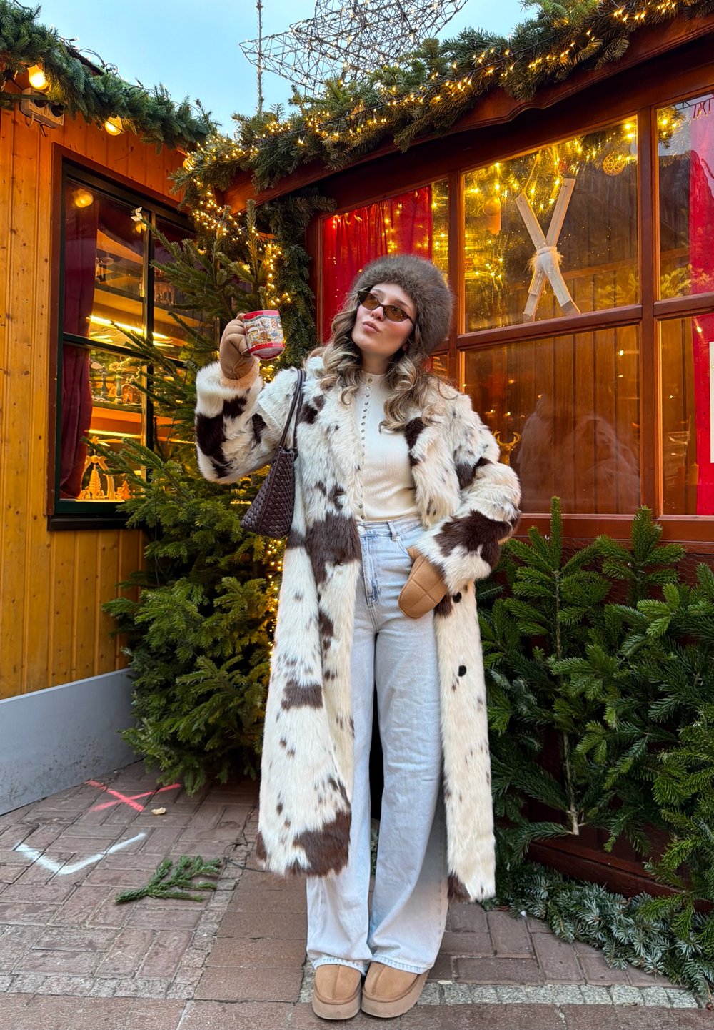 Long manteau en fausse fourrure blanc avec des taches brunes, haut crème boutonné, jeans larges bleu clair, chaussons beige, tenant une tasse festive.