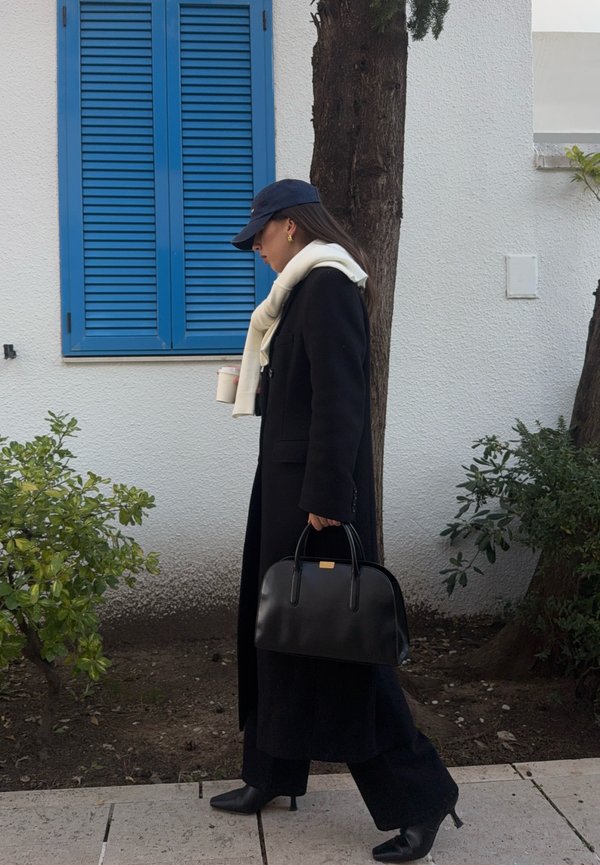 Donna con lunghi capelli scuri che indossa un cappotto nero, una sciarpa bianca e un berretto blu navy che cammina davanti a un edificio bianco con persiane blu, tenendo in mano un caffè e una borsa nera.