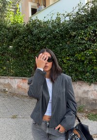 Grijze pinstripe blazer over een witte top, gecombineerd met hoogtaille jeans. Model houdt hand naar gezicht, draagt donkere zonnebril. Zwarte tas met accenten.