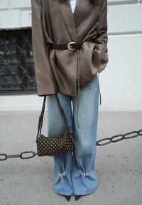 Blazer en satin marron avec taille cintrée, associé à un jean flare bleu clair avec des accents métalliques à l'ourlet et un sac à main marron à pois.