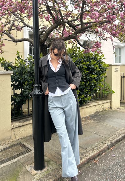 Persona con chaleco gris, camisa blanca, abrigo largo oscuro y pantalones gris claro apoyada en un farol negro bajo un árbol rosado en flor en una calle residencial.