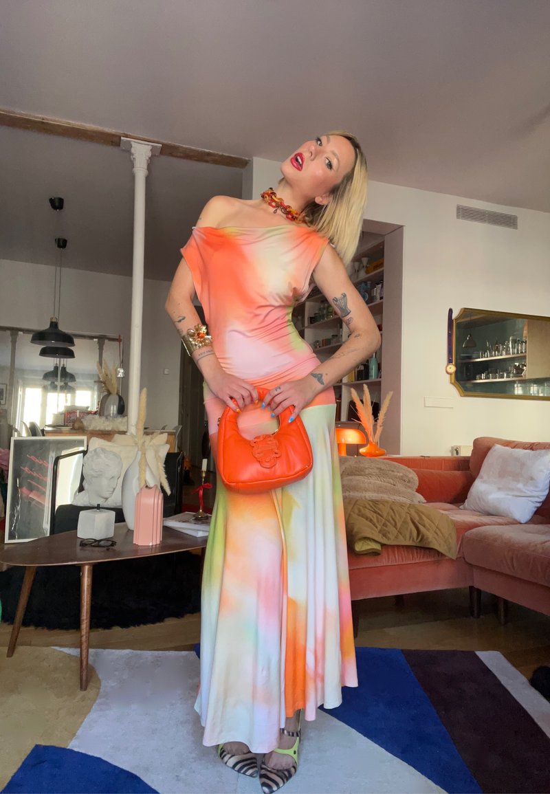 Personne en robe orange à épaules dénudées et tie-dye pastel tenant un sac à main orange vif, debout dans un salon moderne avec une décoration colorée.
