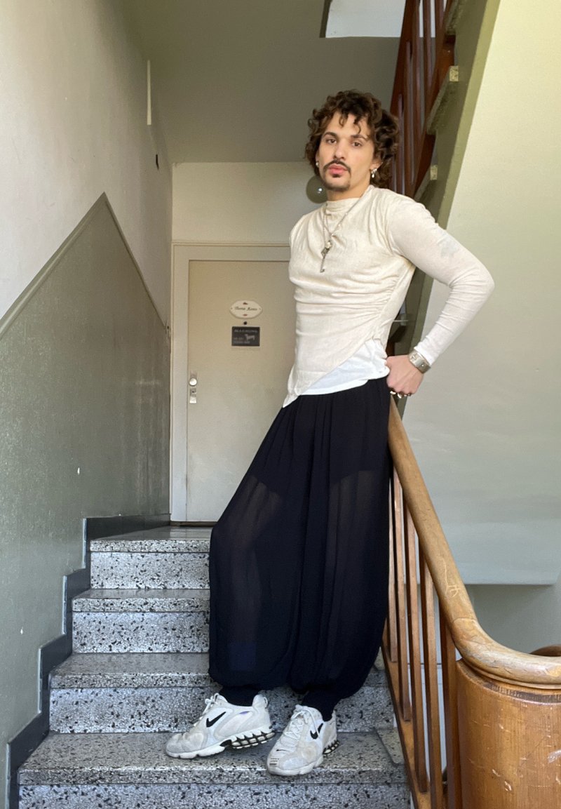 Personne aux cheveux bouclés portant un haut beige à manches longues, un pantalon large noir et des baskets blanches Nike, debout sur un escalier, appuyée sur la rampe.