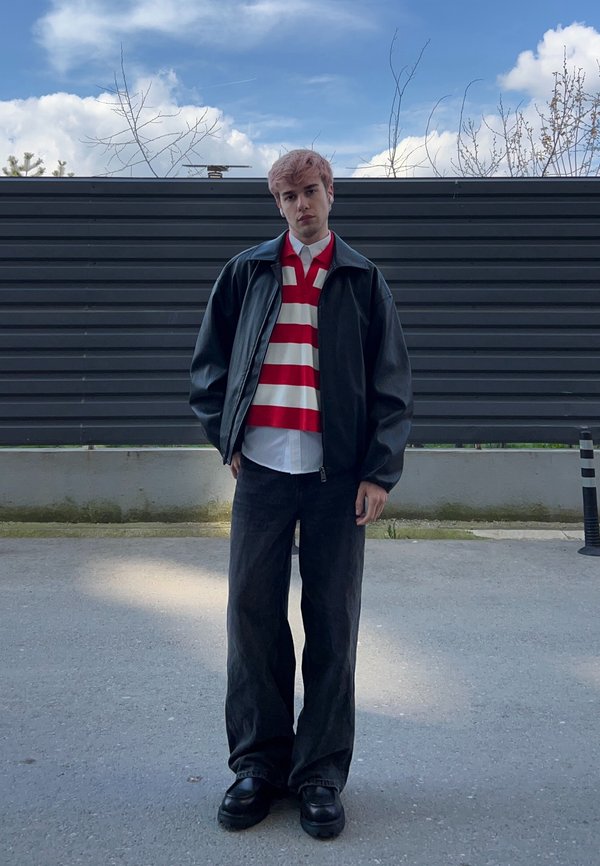 Jeune personne portant un polo rayé rouge et blanc, une veste en cuir noire, un pantalon noir large, debout sur un sol pavé face à une clôture sombre sous un ciel bleu.