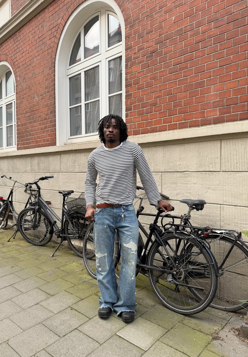 Homme portant une chemise rayée à manches longues et un jean usé tient un vélo noir sur un trottoir à côté d’un bâtiment en briques rouges.
