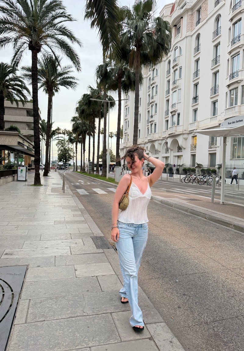 Femme aux cheveux courts et bouclés, portant un haut blanc et un jean bleu, marchant le long d'une rue de la ville bordée de palmiers près de bâtiments blancs.
