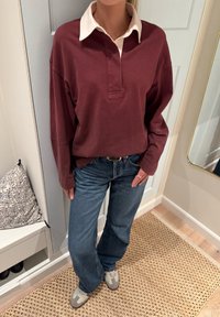 Bordowa koszulka polo z długim rękawem i białym kołnierzykiem, zestawiona z niebieskimi dżinsami i szarymi trampkami. Tkanina wydaje się miękka i casualowa.