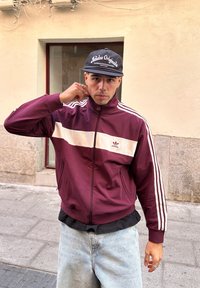 Jachetă de trening maron cu dungi albe și logo adidas, cu fermoar, manșete elastice. Purtați peste o cămașă neagră, asortată cu blugi bleu deschis.