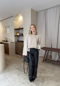 Γυναίκα με μπεζ πουλόβερ και μαύρο δερμάτινο παντελόνι κρατώντας ένα φλιτζάνι και τσάντα, στέκεται σε ένα μινιμαλιστικό δωμ άτιο με ξύλινα έπιπλα και κουρτίνες.