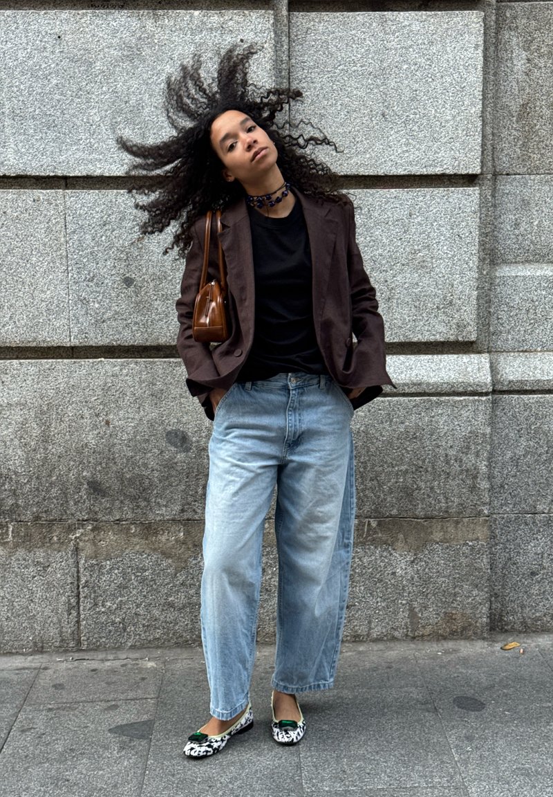 Joven con cabello rizado moviéndolo, lleva puesto un blazer marrón, una camiseta negra, jeans de un azul claro, unos mocasines con estampado animal y un bolso marrón.