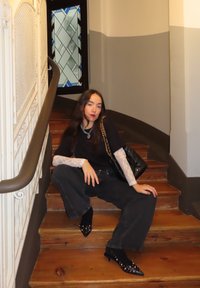 Zwart topje met kanten mouwen, donkere wijde jeans, zwarte puntige schoenen met studs. Zittend op houten trappen met een decoratieve leuning.