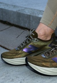 Zöld és arany sportcipők hálós felsőrésszel, lila logóval, fekete-fehér mintás fűzőkkel és párnázott talppal.