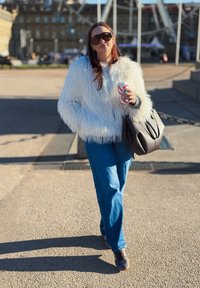Femme portant une veste en fausse fourrure blanche et des lunettes de soleil, tenant une tasse de café et un sac à main noir, marchant à l'extérieur sous le soleil.