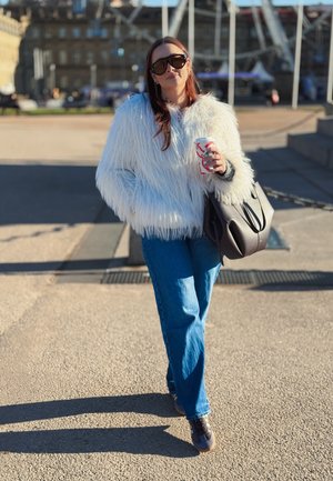 Mujer con chaqueta de piel sintética blanca y gafas de sol, sosteniendo una taza de café y un bolso negro, caminando al aire libre bajo la luz del sol.