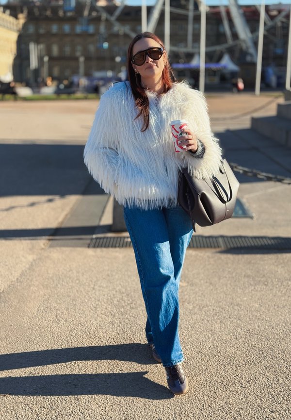 Woman wearing a white faux fur jacket and sunglasses, holding a coffee cup and a black handbag, walking outdoors in sunlight.