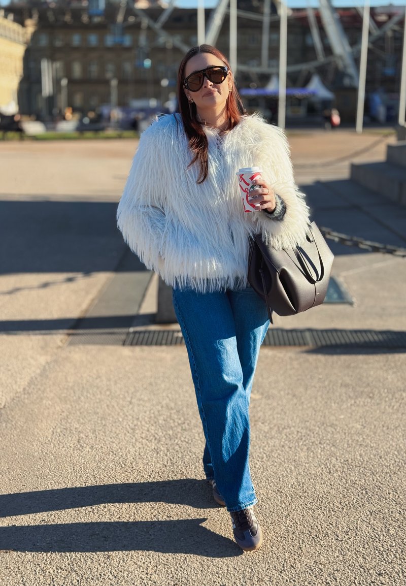 Femme portant une veste en fausse fourrure blanche et des lunettes de soleil, tenant une tasse de café et un sac à main noir, marchant à l'extérieur sous le soleil.