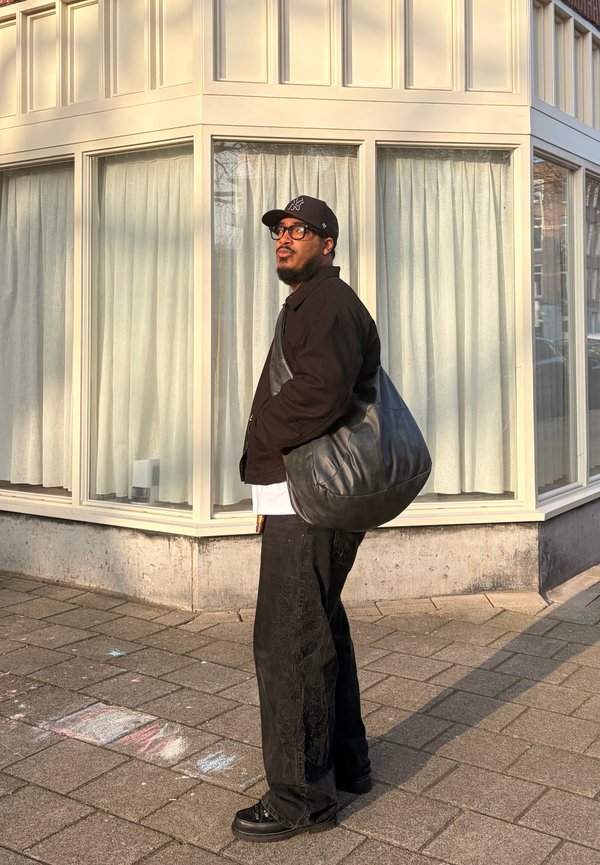 Homme portant une casquette noire, des lunettes, une veste noire et un pantalon, debout sur un trottoir près d'un coin de bâtiment avec de grandes fenêtres et des rideaux blancs.