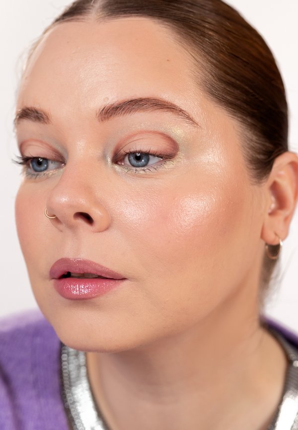 Gros plan sur une femme à la peau claire, aux yeux bleus, au maquillage subtil, portant un anneau de nez, une petite boucle d'oreille et un haut violet avec un col argenté.