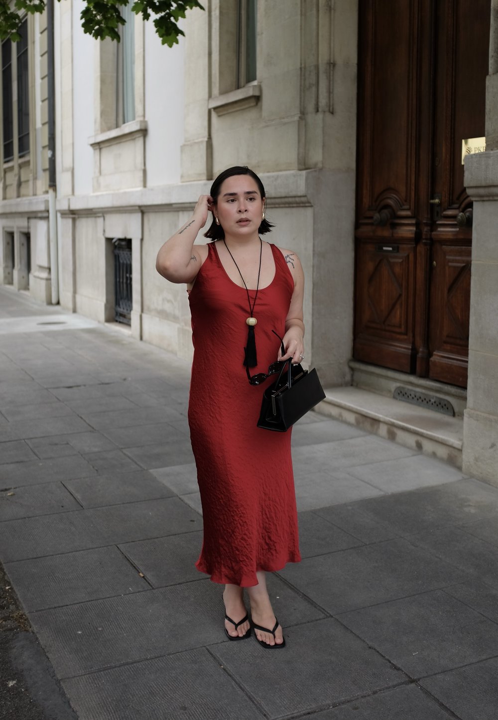 Donna in un vestito rosso senza maniche che cammina su un marciapiede cittadino, con una borsa nera e occhiali da sole, e capelli corti scuri.