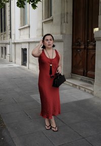 Donna in un vestito rosso senza maniche che cammina su un marciapiede cittadino, con una borsa nera e occhiali da sole, e capelli corti scuri.