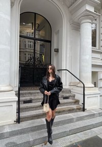 Giacca nera in pelle, vestito nero con rifiniture in pizzo, stivali neri fino al ginocchio, piccola borsa dorata; dietro scalini e porta decorata.