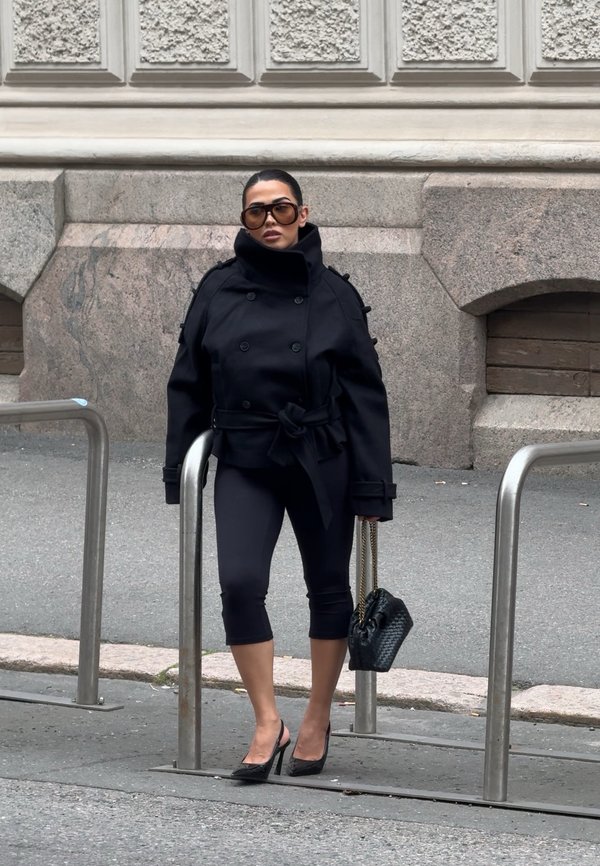 Femme portant une veste noire oversize, un pantalon capri noir, des talons hauts noirs, de grandes lunettes de soleil, tenant un sac à main noir, debout près de rampes métalliques dans la rue.