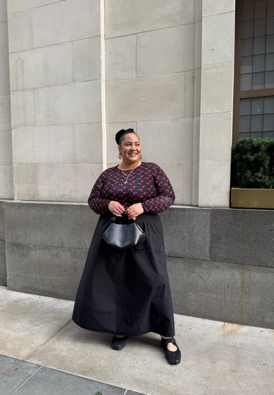 Larga falda negra, blusa de manga larga con patrón de media luna roja, bolso negro y zapatos negros con tiras. De pie contra una pared de piedra.