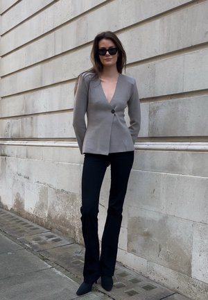 Blazer gris ajustado con escote en V profundo, cierre de un solo botón, combinado con pantalones negros acampanados y zapatos negros de punta frente a una pared de piedra.