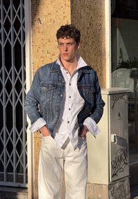 Jeune homme aux cheveux courts et bouclés, portant une chemise blanche et une veste en denim, debout les mains dans les poches contre un mur carrelé au soleil.