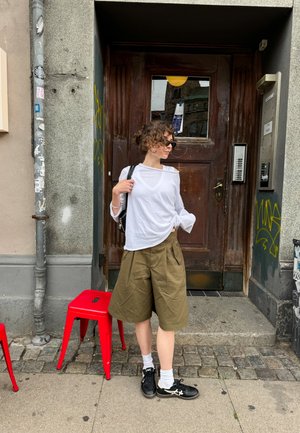 Weißes, leichtes Langarmhemd und olivgrüne, weite Shorts. Schwarze Sneaker mit weißen Akzenten. Rote Metallhocker im Vordergrund.