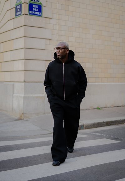 Sudadera negra con cremallera y un bolsillo frontal, combinada con pantalones negros holgados. Presenta gafas transparentes y un peinado gris claro.