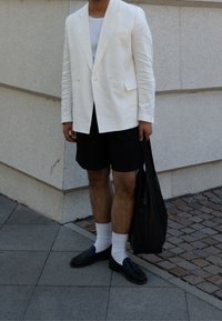 Blazer blanco de lino, camiseta blanca de cuello redondo, pantalones cortos negros, mocasines negros y calcetines blancos. Sosteniendo un bolso tote negro. Entorno al aire libre.