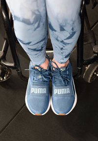 Chaussures de sport en mesh bleu avec tissu texturé, affichant un logo "PUMA" blanc bien visible et des accents noirs. Portées avec des leggings bleus à motifs.