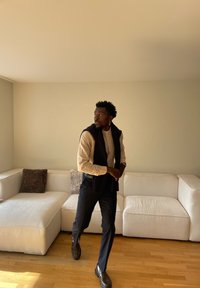 Bărbat în cămașă bej cu pulover negru așezat pe umeri, pantaloni negri și mocasini, mergând într-o cameră cu o canapea albă și podea din lemn.