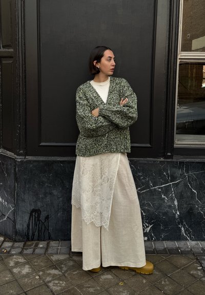 Cárdigan verde de punto con mangas oversized, superpuesto a un vestido crema con encaje. Presenta zapatos de plataforma amarillos y un fondo texturizado.