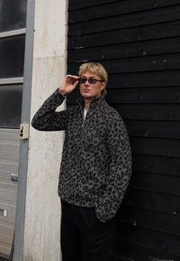 Pulover iz flisa z leopardjim vzorcem v sivi barvi in visokim ovratnikom. Model pozira ob črni leseni steni, nosi očala in črne hlače.