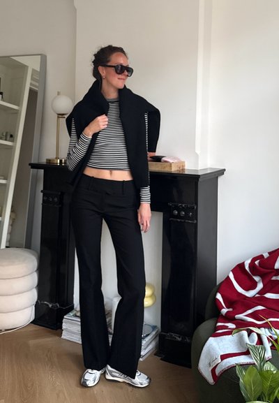 Mujer con gafas de sol negras, pantalones negros, top corto de rayas y suéter negro sobre los hombros, de pie en una sala de estar cerca de una chimenea.