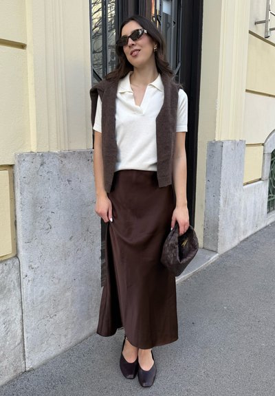 Camisa de polo blanca, chaleco de punto marrón, falda maxi de satén marrón y zapatos de tacón marrón oscuro. Bolso de mano de tejido en color a juego.