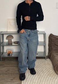 Chemise henley noire à manches longues côtelée, jean denim bleu clair à jambes larges, et chaussures noires à semelle épaisse, sur un fond neutre.