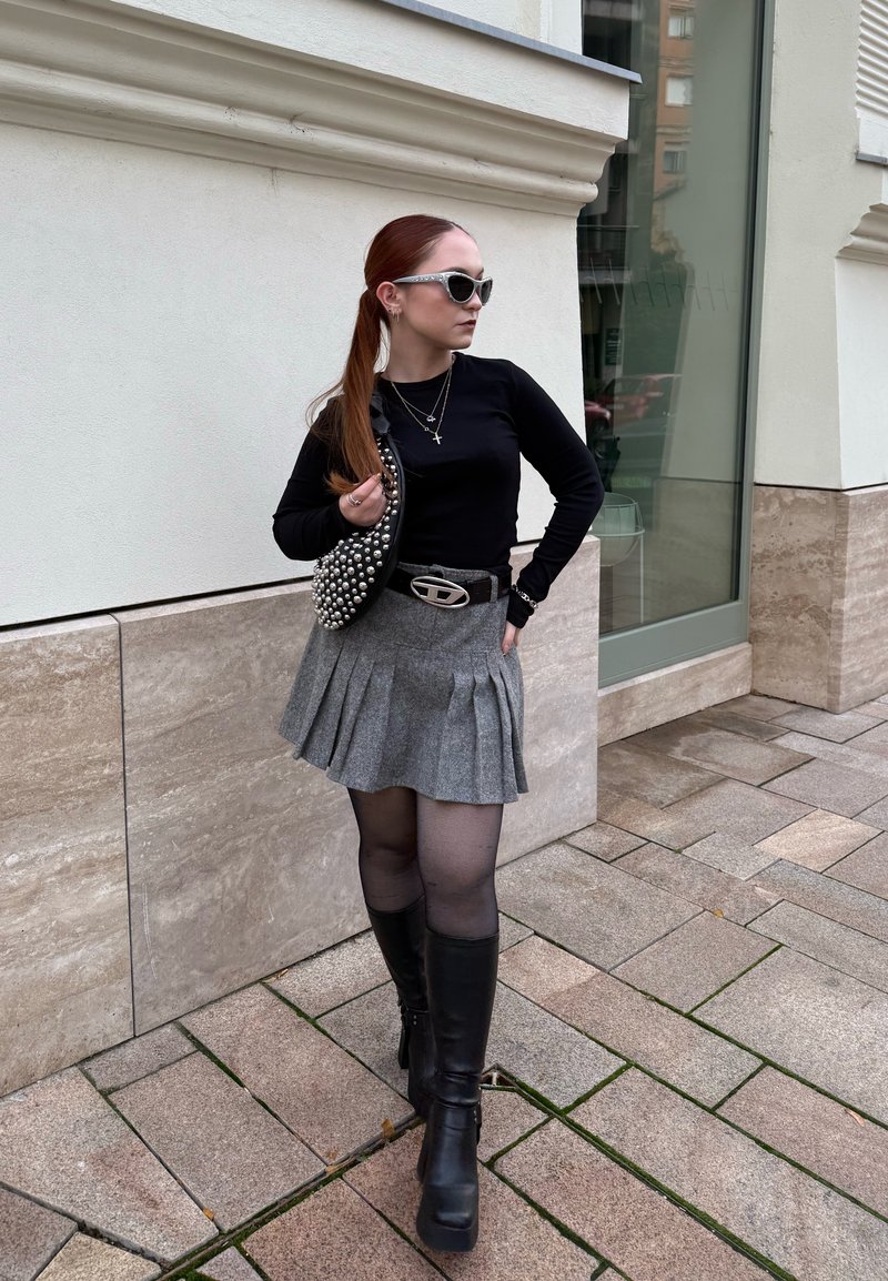 Haut noir à manches longues, jupe plissée grise, bottes noires montantes jusqu'aux genoux, ceinture argentée, sac à main orné et lunettes de soleil posés devant un mur clair.