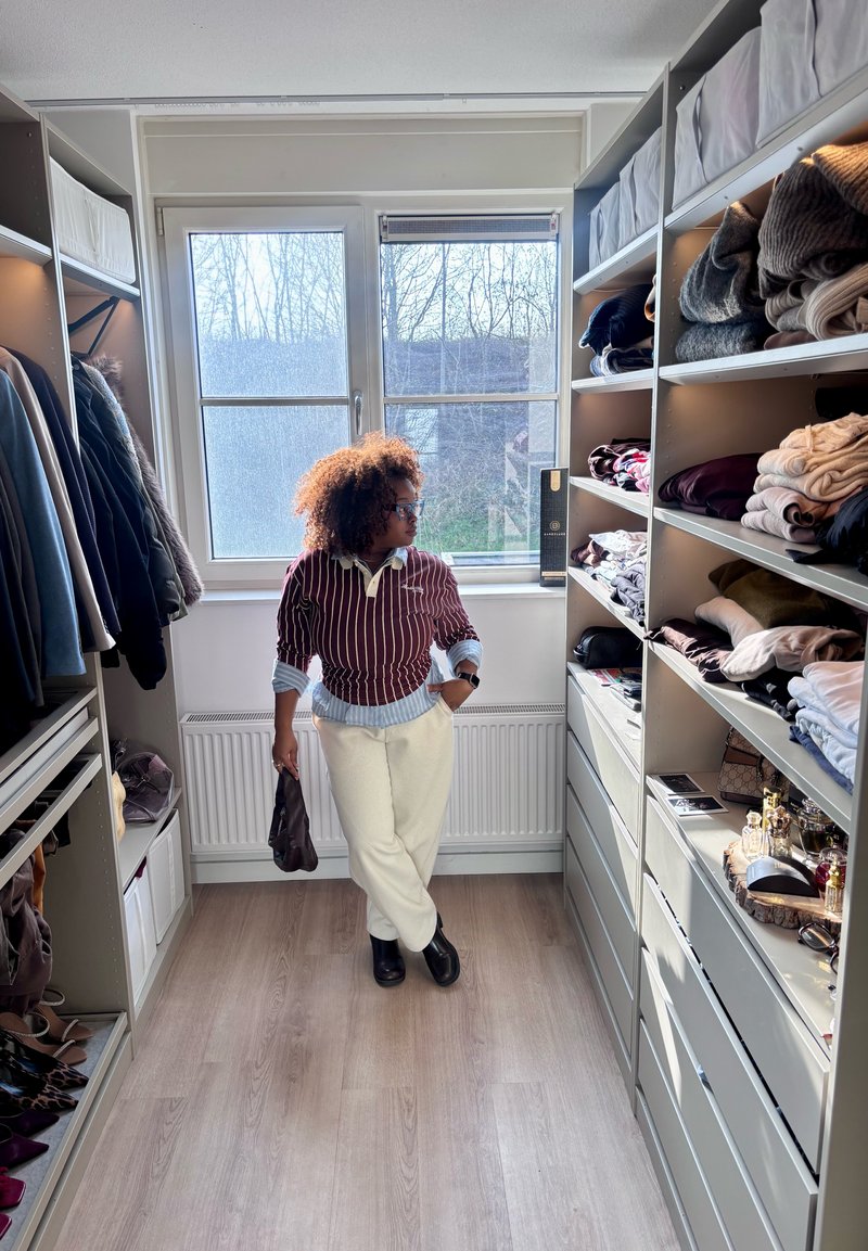 Osoba s kovrčavom kosom drži torbu i stoji u dobro osvijetljenom garderobnom ormaru s policama punim složene odjeće i vješalicama s kaputima.