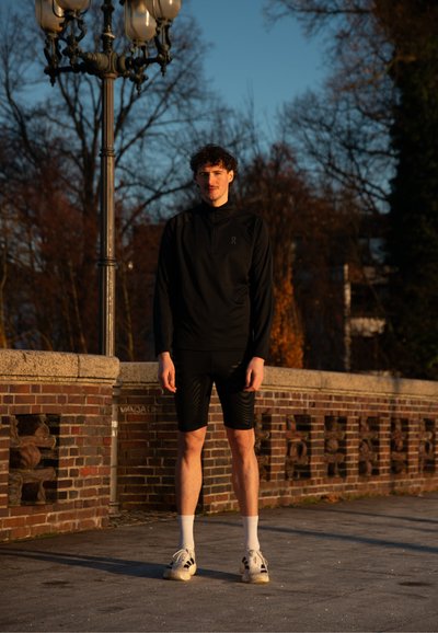 Joven con ropa deportiva negra y zapatillas blancas junto a una pared de ladrillos bajo una farola alta al atardecer.
