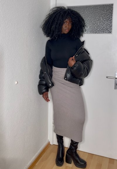 Chaqueta de plumas negra, falda larga de punto gris con canalé, botas negras de tacón altas hasta la rodilla. Texturas suaves; blusa ajustada, de pie contra una pared clara.