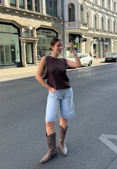 Top de punto sin mangas marrón, pantalones cortos de mezclilla clara y botas de vaquero marrones oscuras con diseños bordados, posado en una calle urbana.