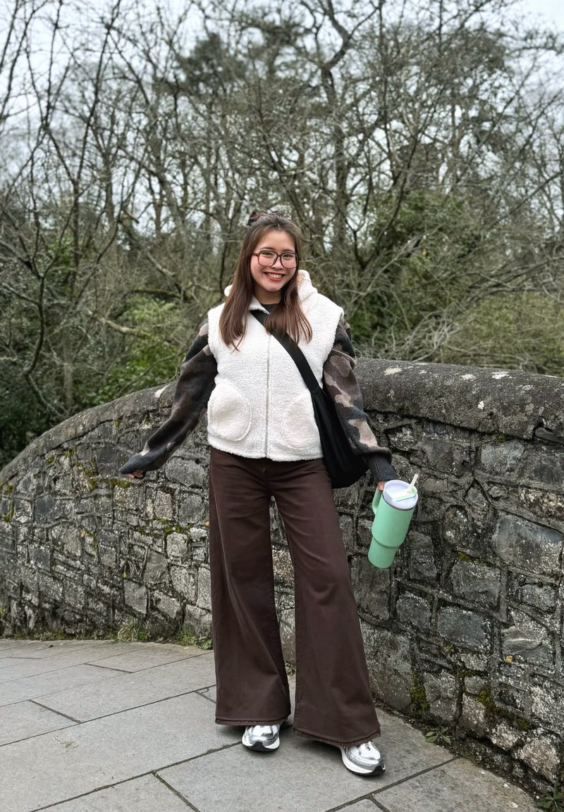 Jeune femme en lunettes, gilet en polaire blanc, manches camouflage et pantalon large marron, souriante, debout près d’un pont en pierre en tenant un gobelet vert.