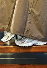 Γκρι και άσπρα αθλητικά παπούτσια με δίχτυ και συνθετικά υλικά, τοποθετημένα σε ξύλινη επιφάνεια, εν μέρει καλυμμένα από ένα καφέ, ρέον ύφασμα.